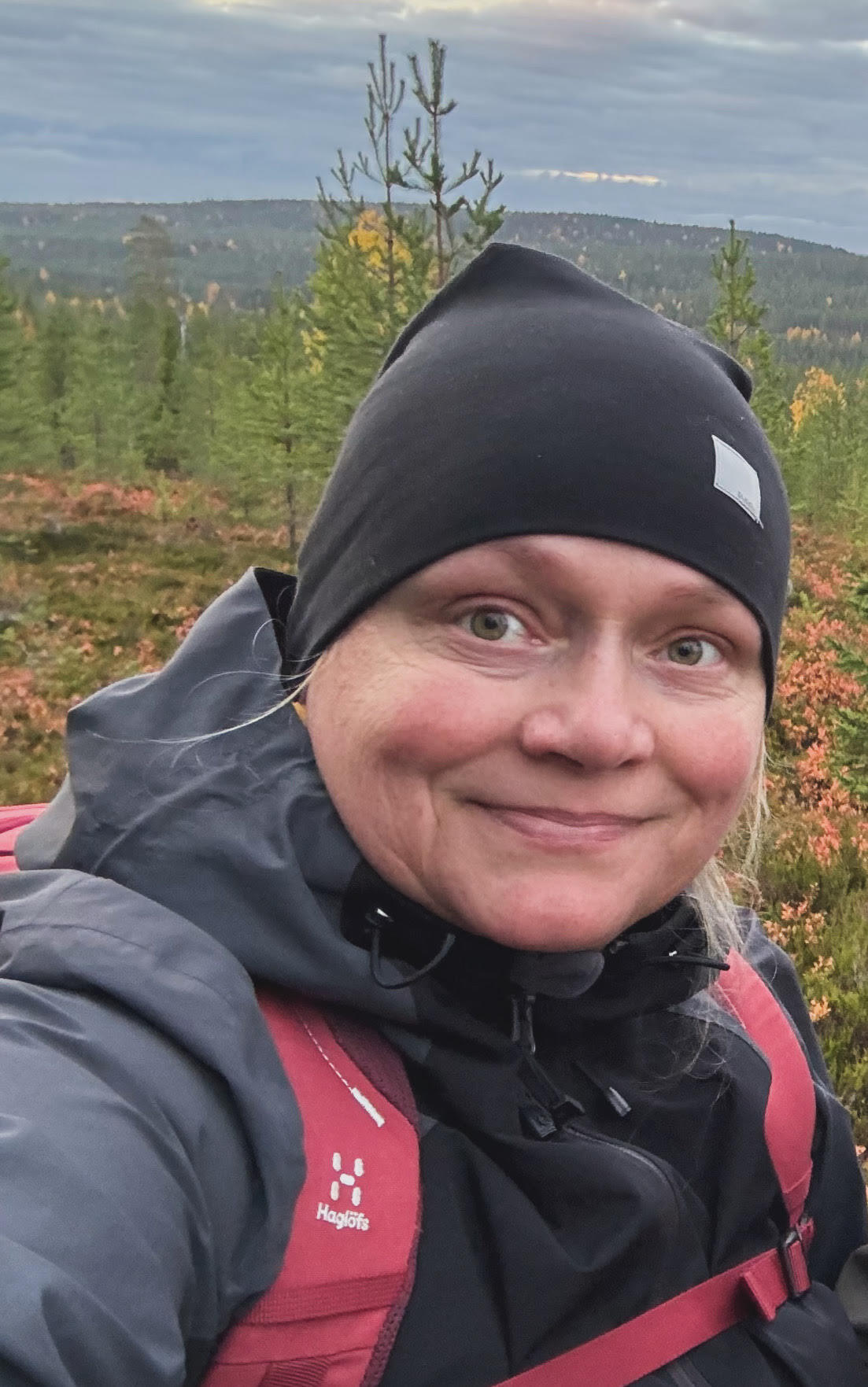 Nainen katsoo kameraan ja hymyilee lempeästi. Hänellä on ulkovaatteet päällä ja taustalla on metsää.
