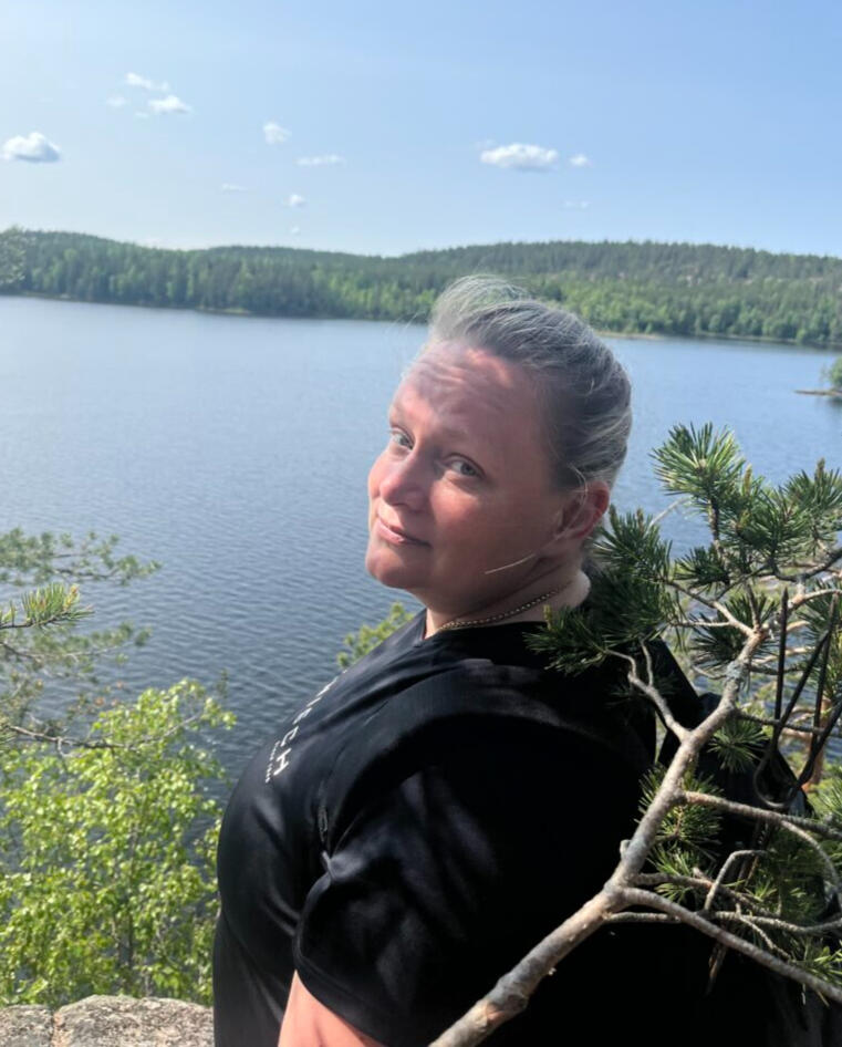 Nainen seisoo metsässä järven edessä. Hän on kääntänyt katseen kameraan ja hymyilee tyynesti.